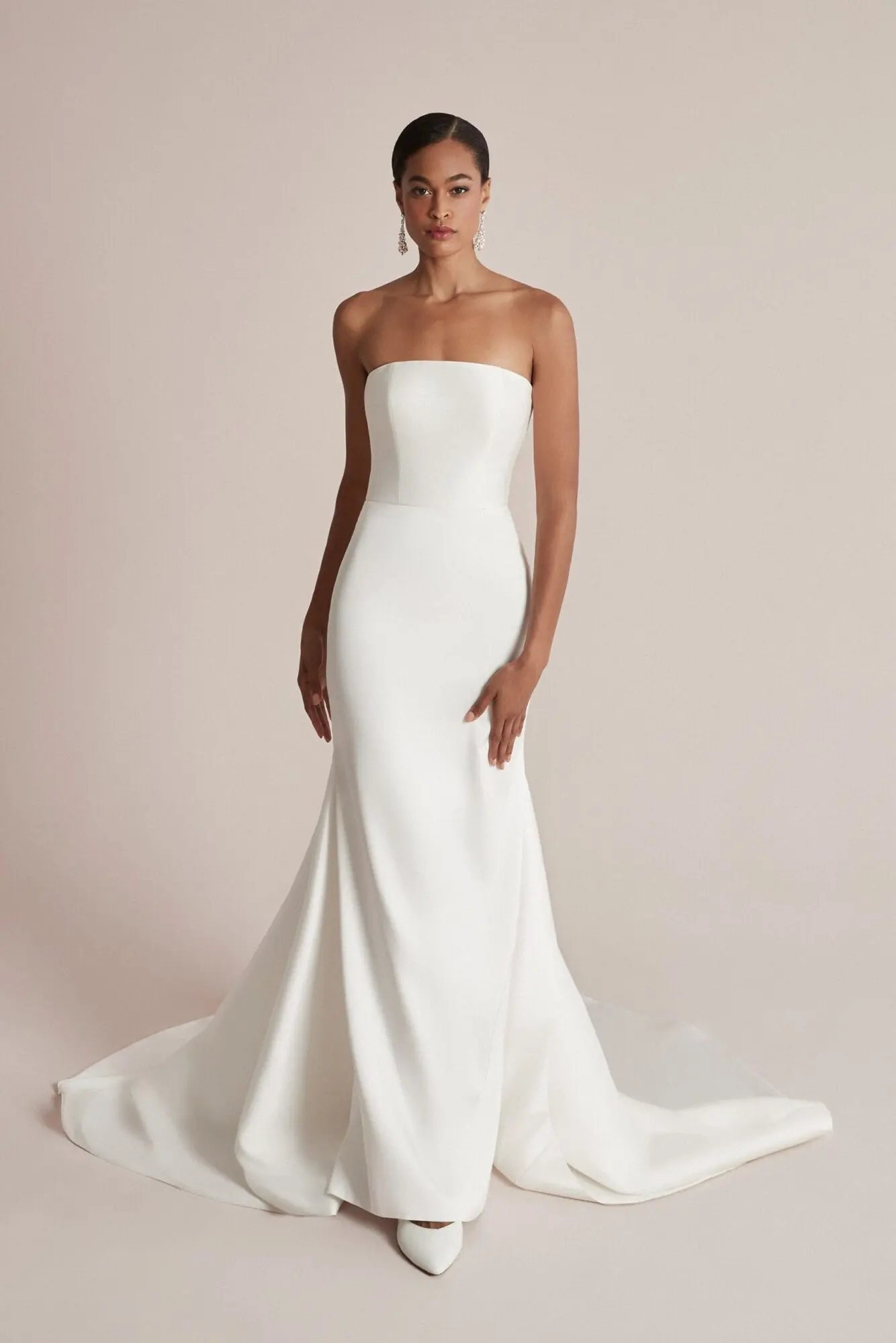 1711226026381-img-1973-2 Celeste, an ivory stretch Mikado fit-and-flare bridal gown by Justin Alexander, featuring a chic straight strapless neckline, princess seams along the bodice, and elegant inverted godets that enhance the skirt's chapel train. The dress is modeled by a woman against a light, plain background.