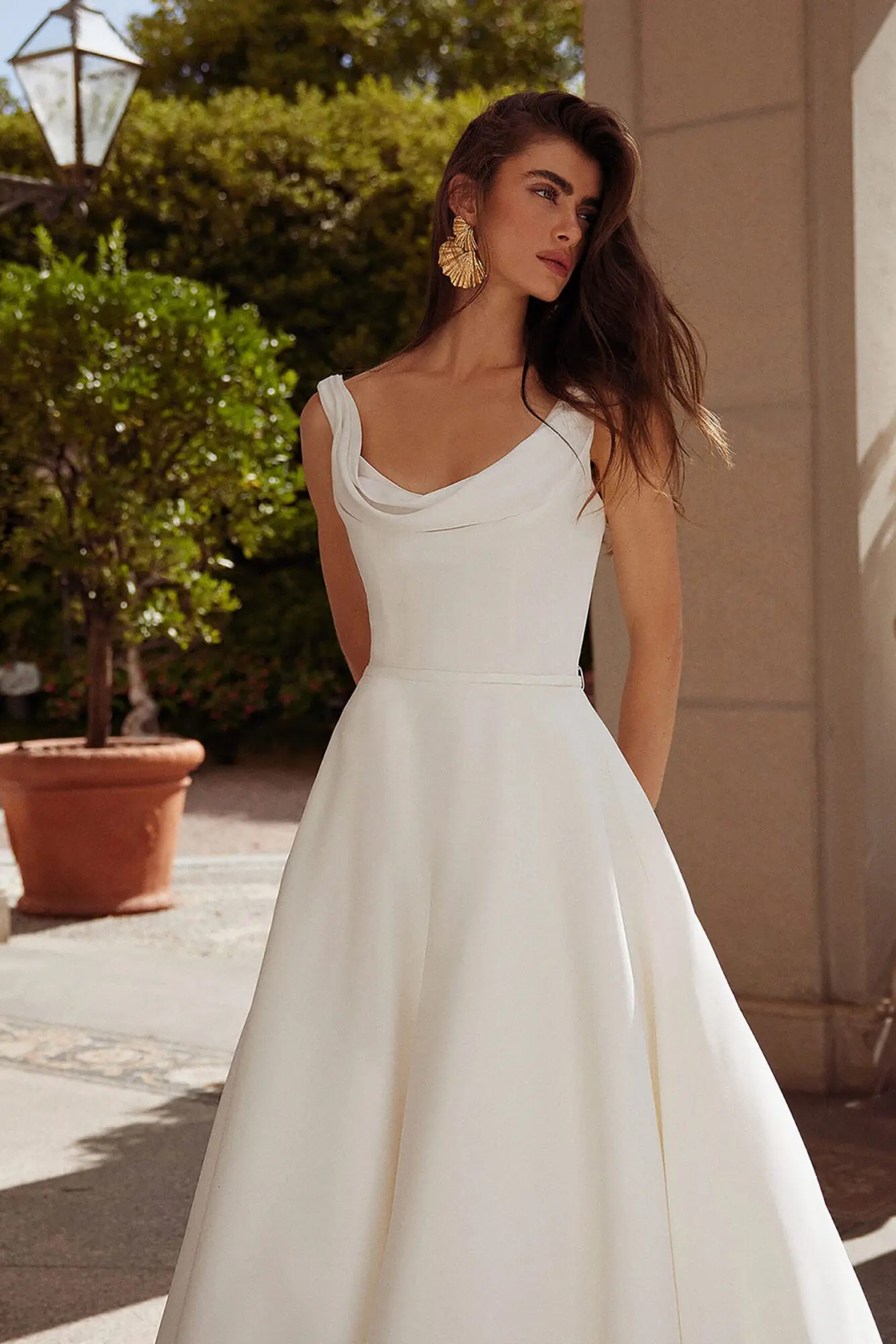 1711227220861-img-1974-1 Grove, a Justin Alexander off-white/ivory bridal ball gown with a draped cowl neckline and a clean crepe bodice. The gown features a full satin skirt, a thin detachable satin belt, discreet pockets, and a chapel-length train with satin-covered buttons. A model with long dark hair and golden earrings poses outdoors in sunlight.