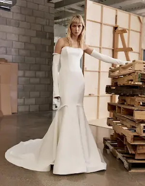 1715701747837-nola1-2 Nola, an ivory strapless fit-and-flare satin bridal gown with a boned bodice, belled detachable arm sleeves, and a chapel train, worn by a blonde model in an industrial-style space with a brick wall and stacked wooden pallets.