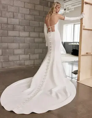 1715701747838-Nola2-1 Nola, an ivory satin strapless fit-and-flare bridal gown by Alyne, features a boned bodice with a corset lace-up back and a chapel train adorned with a row of delicate bows. The model wears long, fitted ivory gloves.