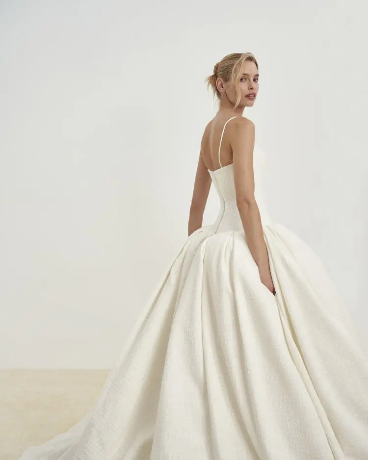 1729784081931-image-15-2 Senstudio SE048, an ivory tweed drop-waist ballgown with delicate spaghetti straps and a voluminous pleated skirt. A model wearing the gown looks over her right shoulder, showcasing its textured fabric and elegant silhouette.
