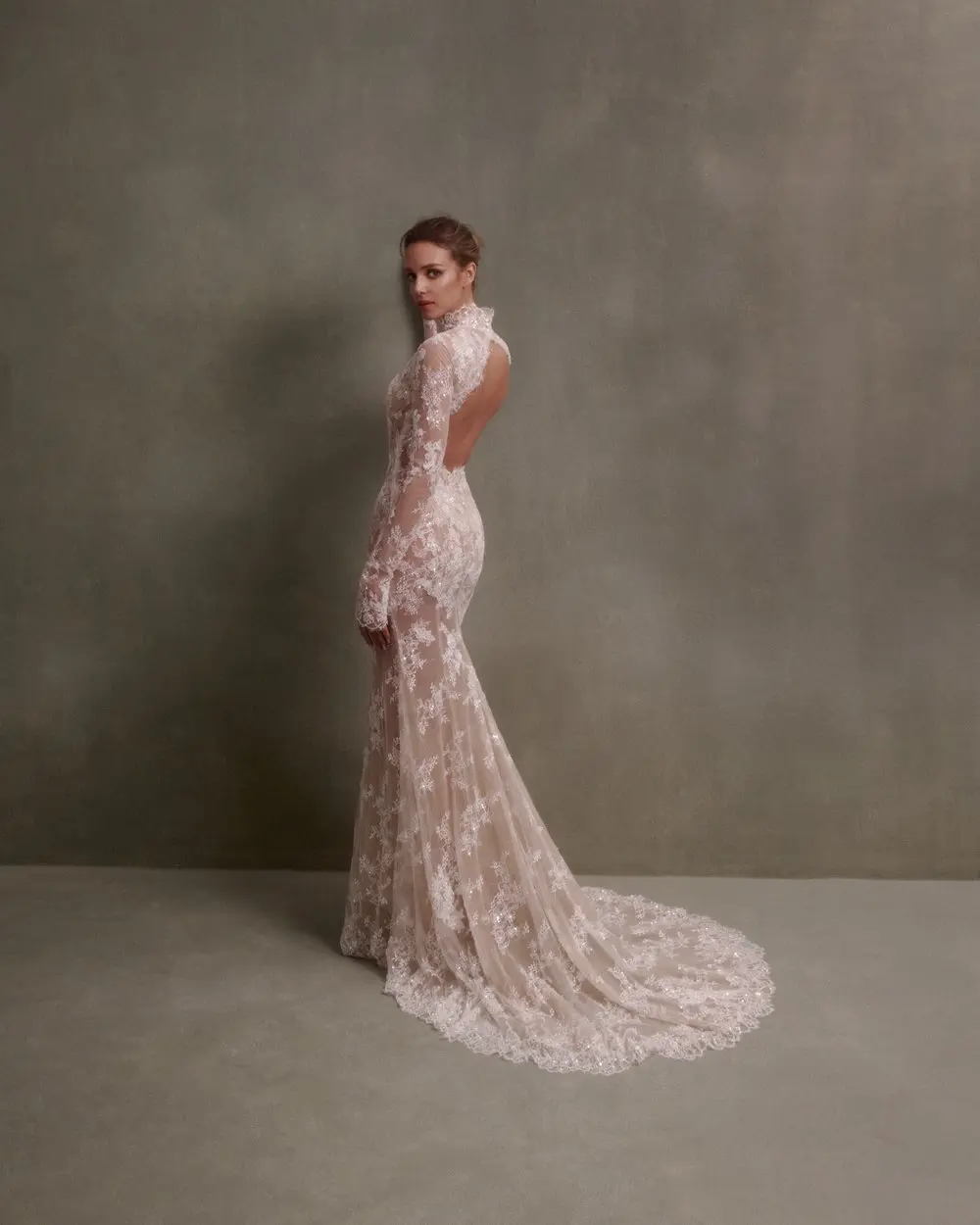 1730138478011-image-1-2 NF082, an ivory and nude mermaid bridal gown by Nicole + Felicia. A model stands in profile, turning her head to look directly at the viewer, showcasing the stunning long-sleeve gown against a textured gray-green wall. The dress features a high neckline, a dramatic open back, intricate sheer lace detailing with beading, and a flowing chapel train.