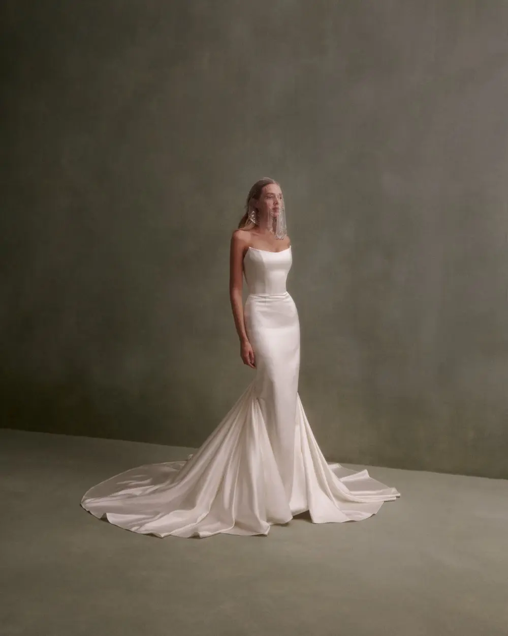 1730139106651-image-2-2 NF086, a cream satin mermaid strapless gown by Nicole + Felicia, featuring a soft scoop neckline and a dramatic pleated train, worn by a model against a textured green background.