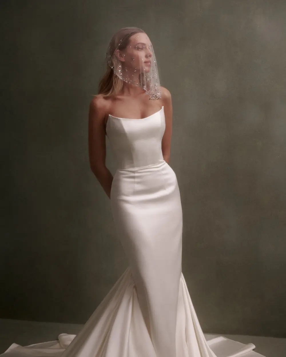 1730139106652-image-3-2 NF086, a cream Nicole + Felicia satin mermaid strapless wedding gown featuring a soft scoop neckline and a dramatic pleated train. A blonde woman models the elegant gown, looking to the side with her hands clasped behind her back, while wearing a sparkly birdcage veil against a muted green background.