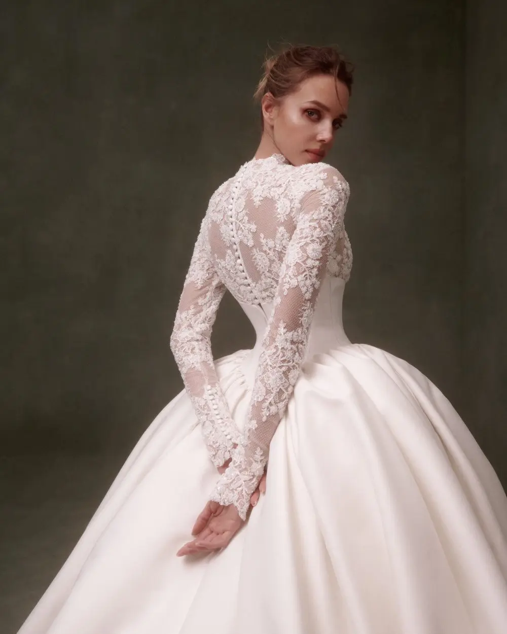 1730139233346-image-5-2 NF088, an ivory and nude long-sleeve ballgown wedding dress, featuring a high-necked lace bodice with intricate button details down the back and delicate lace sleeves, flowing into a voluminous mikado skirt. The model looks over her left shoulder, with her hands clasped in front of her.