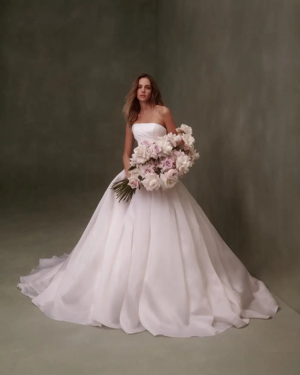 1730139393511-image-7-5 A woman models a Nicole + Felicia Silk White strapless silk organza drop-waist bridal gown with a voluminous, ethereal ball gown skirt and a long train, holding a large bouquet of pastel pink and white roses against a dark green studio backdrop.