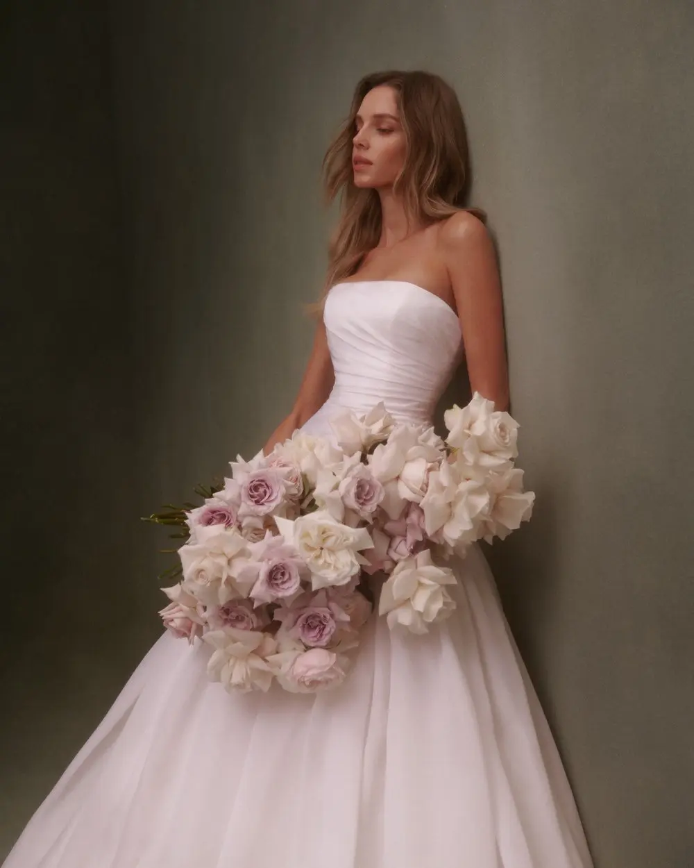 1730139393512-image-8-2 NF089, a Silk White silk organza strapless drop-waist ball gown, showcases modern allure and soft romance with its elegant bodice and flowing, ethereal skirt. A model with light brown hair leans against a muted green wall, holding a large bouquet of pastel pink, lavender, and cream roses.