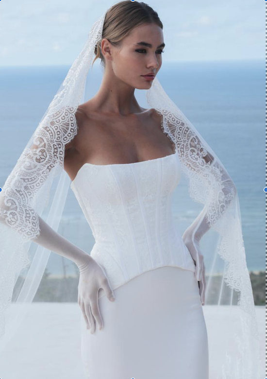 1732120140898-thea-bodice-wedding-dress-2 Tori Skirt, an ivory satin bridal separates gown by Mariana Hardwick, featuring a sleek, body-hugging slim A-line Tori skirt and a structured strapless Thea bodice with subtle lace. The model completes the look with a long, intricate lace-trimmed veil and elegant sheer elbow-length gloves, posing gracefully against a bright blue ocean backdrop.