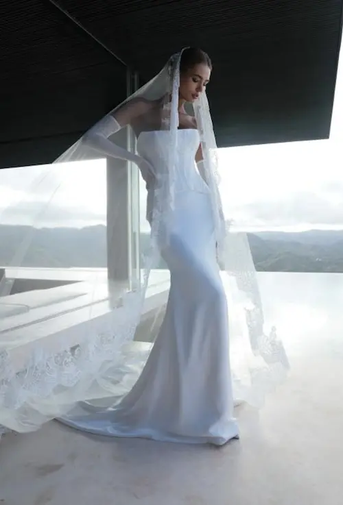 1732120140899-thea-bodice-wedding-dress-2-5 Mariana Hardwick's 'Tori Skirt' and 'Thea Bodice' ensemble, an ivory strapless satin slim A-line bridal gown, consisting of a structured corset bodice and a sleek, flowing skirt. The look is completed with a long, lace-trimmed veil and elegant elbow-length gloves. A model stands gracefully indoors by a large window overlooking a scenic mountain and ocean landscape.