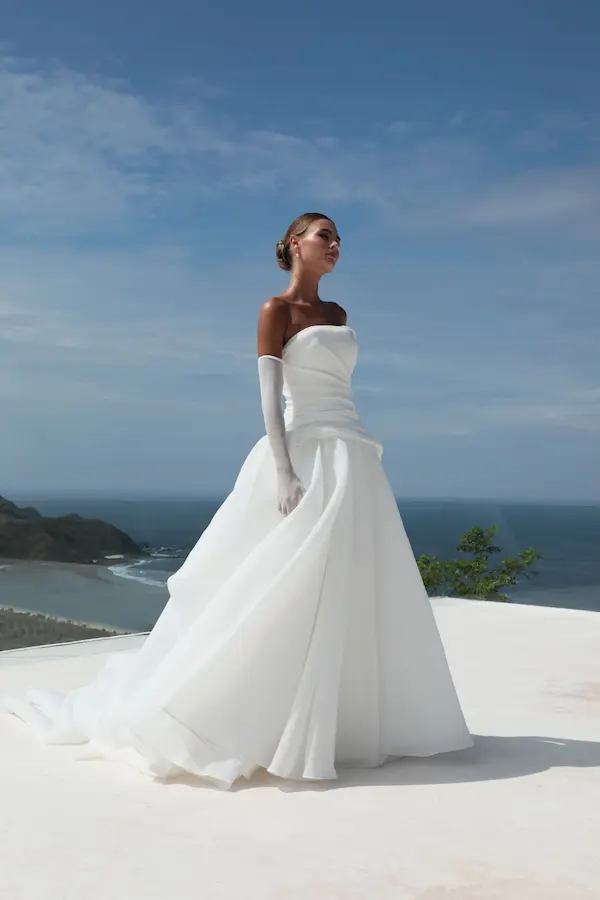 1732125831167-alison-1-5 Alison, an ivory strapless organza ball gown by Mariana Hardwick, features a beautifully draped drop waist and a voluminous, airy silhouette. The bride, with her hair in a bun and wearing long white gloves, stands elegantly on a light-colored outdoor terrace overlooking a scenic ocean bay under a bright blue sky.