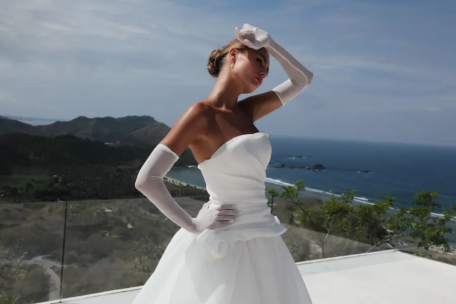 1732125831168-alison-2-2 Alison, an ivory strapless organza ball gown by Mariana Hardwick, featuring a beautifully draped drop waist and handmade floral motifs. A bride wearing the voluminous gown and long white sheer gloves poses on a sunlit balcony, shielding her eyes with one hand. The background reveals a stunning coastal landscape with mountains, a beach, and the expansive ocean under a clear sky.