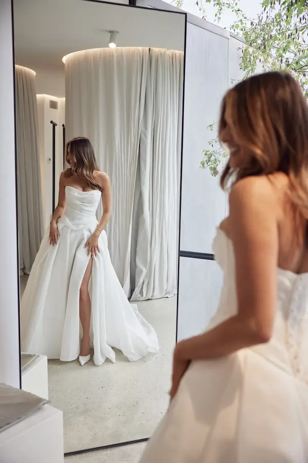 1732126471723-valencia-5-2 Valencia, an ivory, strapless A-line bridal gown by Mariana Hardwick, featuring a draped organza demi-sheer bodice with intricate corset detailing and a dramatic pleated skirt with a high slit and cathedral train. A smiling woman tries on the gown in a fitting room, admiring her reflection in a full-length mirror while wearing white pointed-toe heels.