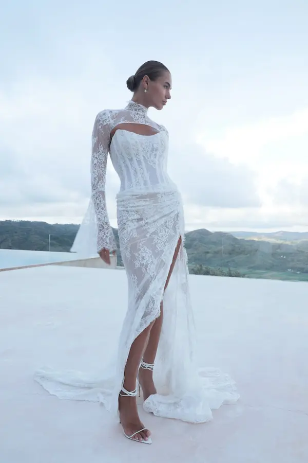1732127260044-alia-1-2 Alia, a white Mariana Hardwick bridal gown featuring a draped and corseted lace fit-and-flare silhouette with a thigh-high slit, complemented by a sheer lace bolero with a high neck and flared sleeves. The elegant ensemble is worn by a model with a sleek bun and pearl earrings, standing on a bright white outdoor terrace overlooking distant mountains.