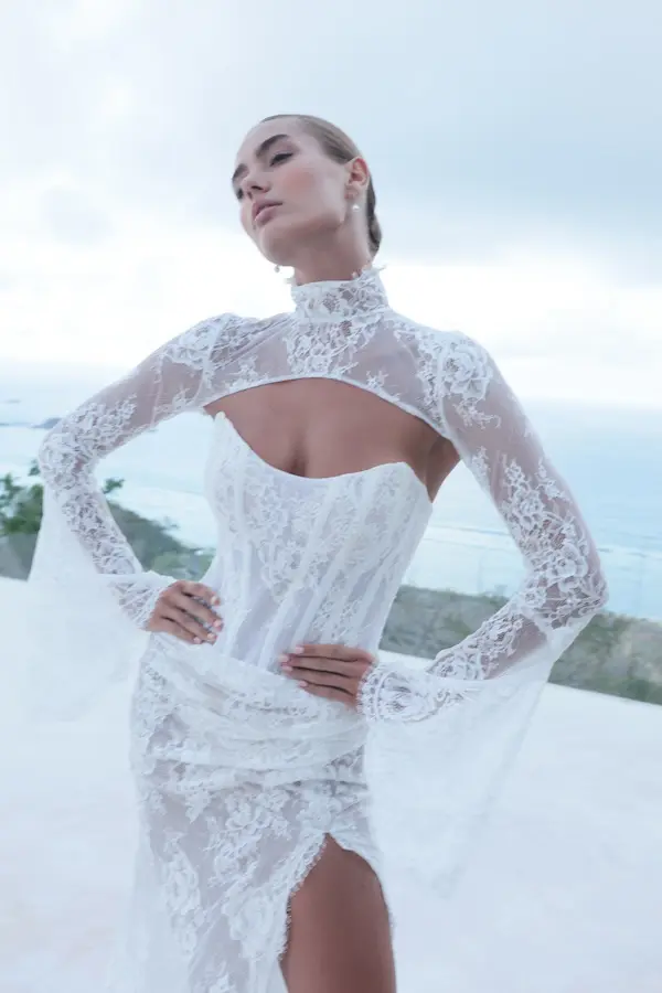 1732127260046-alia-w-lace-bolero-3-2 Alia, an as-sampled lace fit-and-flare bridal gown with a corseted, strapless bodice and a thigh-high slit, paired with a matching high-neck lace bolero featuring long bell sleeves. A woman models the sheer lace gown confidently outdoors against a cloudy sky and distant ocean view.