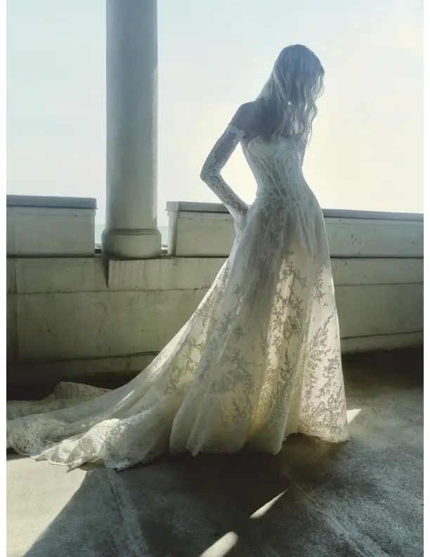 1732640492362-dahlia-1-5 Dahlia, an ivory Basque waist corset A-line gown, features glistening corded lace and detachable lace-appliquéd sleeves worn off-the-shoulder. A model stands backlit on a sunlit stone balcony overlooking the ocean, looking down as the dress's cathedral-length train pools around her.