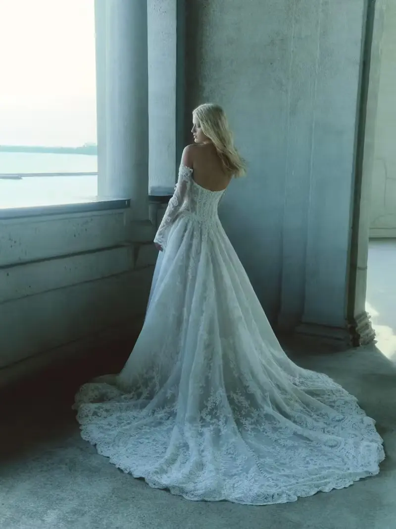 1732640530040-dahlia-2-2 Dahlia, an ivory Basque waist corset A-line gown by Rivini, crafted from glistening corded lace, features a lace button-down back and detachable lace-appliquéd sleeves. A blonde bride models the gown from behind, looking over her shoulder towards a window, showcasing its dramatic cathedral train and intricate lace details.