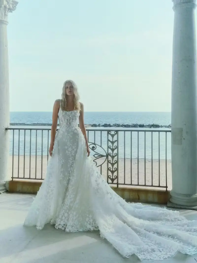 1732640755555-blossom-1-5 Rivini's 'Blossom with Overskirt,' an enchanting ivory 3D laser-cut floral lace corset sheath bridal gown, features a ballerina tank neckline and spaghetti straps, paired with a magnificent detachable ballgown overskirt that creates a long, elegant train. A blonde model poses in the gown on a coastal balcony with the ocean and a sandy beach in the background.