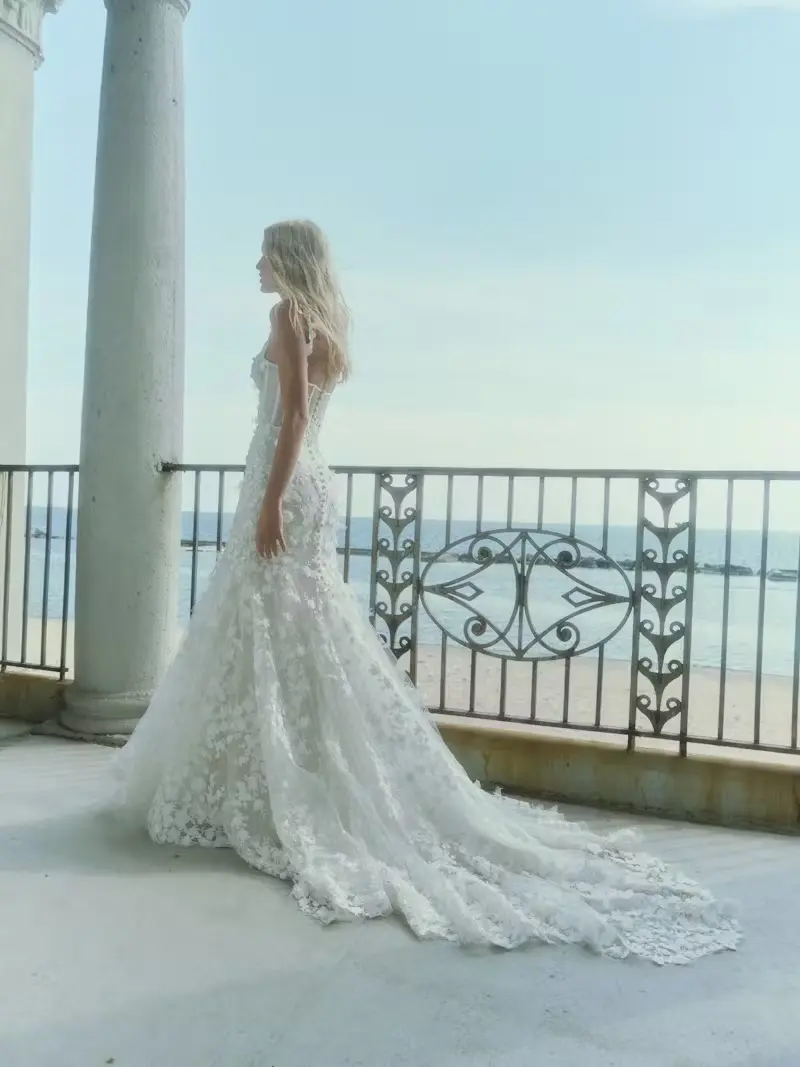 1732640755557-blossom-2-2 Blossom with Overskirt, an ivory Rivini sheath bridal gown featuring a corset bodice, ballerina tank neckline, and spaghetti straps. The dress is adorned with 3D laser-cut floral lace and hand-appliquéd flowers, leading into a dramatic, flowing train, and features a sheer back bodice with a row of buttons. A model with blonde hair stands on a sunlit balcony overlooking the ocean.