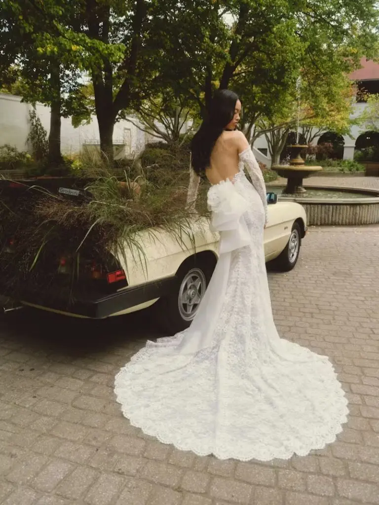 Daisy, an ivory and nude Chantilly corded lace halter sheath gown featuring a striking low back, delicate lace sleeves, and a handmade organza flower adorning a cascading bow that flows into a chapel-length train finished with covered buttons. The bride stands elegantly on a cobblestone path, looking back over her shoulder, with a vintage car filled with lush foliage beside her and a fountain visible in the garden background.