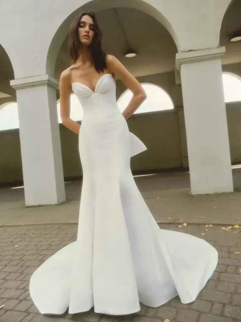 Jasmine, an ivory soft satin fit-and-flare bridal gown with a cathedral train, featuring a strapless draped cat-eye sweetheart neckline, a structured bodice, and a flattering Basque waist. The gown is detailed with a sheer back adorned with boning and delicate buttons trailing to the hem, completed by a dramatic double bow. The model poses elegantly under white archways.