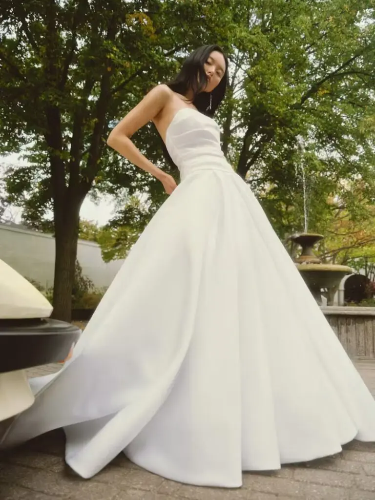 Alyssa, an off-white strapless drop-waist ballgown by Alyne, features a beautifully draped bodice and a voluminous pleated skirt. A model with long dark hair poses with her hand on her hip, looking at the viewer, set against a backdrop of green trees and a stone fountain outdoors.