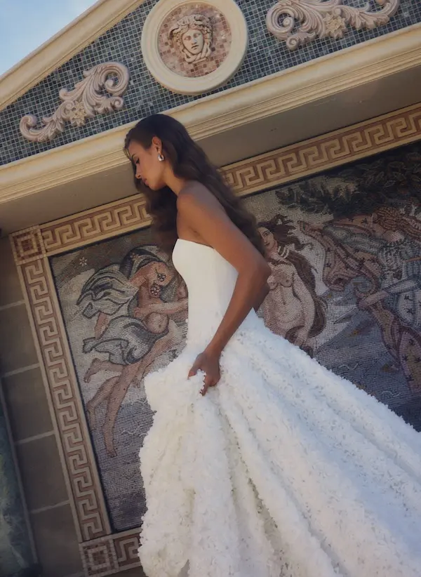 1738945565552-bijou-ldg2-1 Bijou, a white couture wedding gown by Leah Da Gloria. A model with long, flowing brown hair gently looks down as she walks, wearing a strapless white wedding gown featuring a sleek bodice and a voluminous ball gown skirt richly adorned with intricate hand-sewn 3D floral appliqués. She lightly holds a part of the elegant skirt as she passes a grand, classical-inspired building facade, which includes a detailed mosaic wall depicting mythological figures, a Greek key pattern, and a prominent round relief carving of Medusa's head.