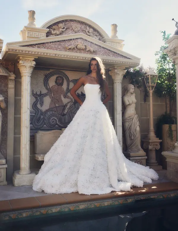 1738945565552-bijou-ldg3-4 Bijou, a white strapless ball gown wedding dress with a dropped waist and intricate 3D floral appliqués adorning its voluminous skirt and long cathedral train. The dress is modeled outdoors by a woman with long brown hair, standing in front of an ornate classical building featuring columns, archways, and a mosaic depicting a figure on a chariot. To her right, a classical statue stands beside a decorative lamp post. The scene is bright and grand, with a pool in the foreground. Designed by Leah Da Gloria.