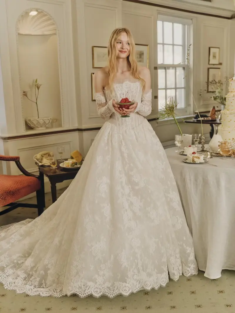 1749147134186-rv-ss26-alyne-dawes-2-front-1 Dawes, an ivory strapless lace Basque waist ballgown by Alyne, is worn by a smiling blonde model. The bridal gown features a demi-sheer lace bodice with micro-pleated tulle at the neckline, a dramatic diaphanous lace skirt with a cathedral-length train and buttons trailing down the back, and elegant removable lace opera cuffs. She holds a small bowl of cherries and stands in an elegant room with a tiered cake and refreshments.