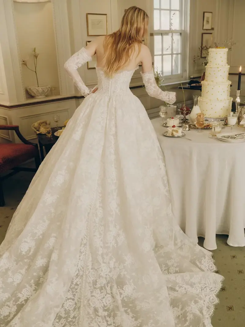1749147134186-rv-ss26-alyne-dawes-3-back Dawes, an ivory strapless lace Basque waist ballgown by Alyne, featuring a translucent bodice, micro-pleated tulle neckline, and a diaphanous lace skirt with subtle volume, flowing into a dramatic cathedral train. The gown is detailed with buttons down the back and styled with elegant lace opera cuffs. The bride, seen from behind, stands beside a dessert table with a tiered wedding cake in a formal room.
