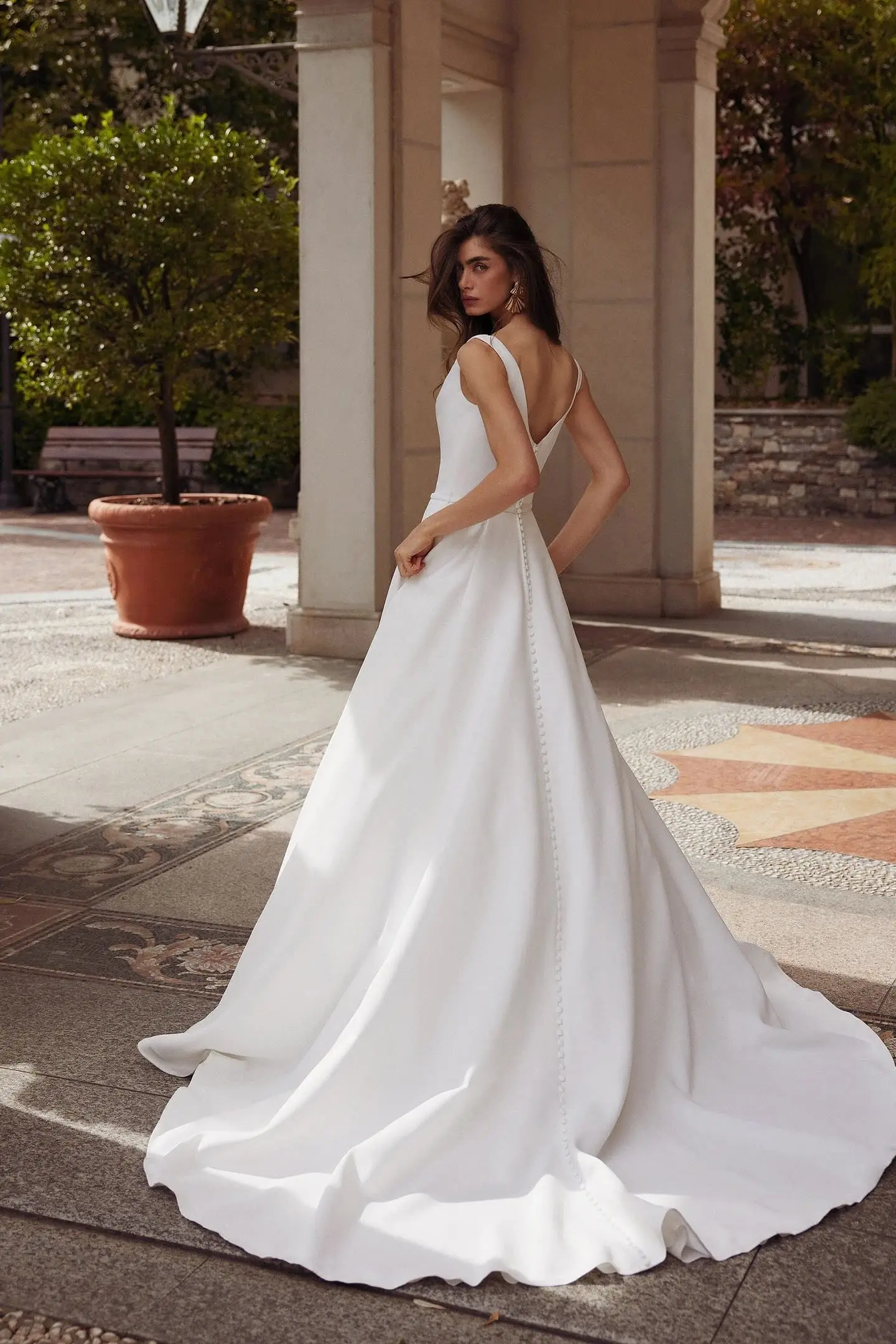 1756498633795-88321_FB_Grove_Justin-Alexander_2-2 Grove, an off-white/ivory Justin Alexander ball gown, features a clean crepe bodice with a deep V-back and a full satin skirt with a chapel-length train adorned with satin-covered buttons, worn by a model looking back over her shoulder in an outdoor courtyard.