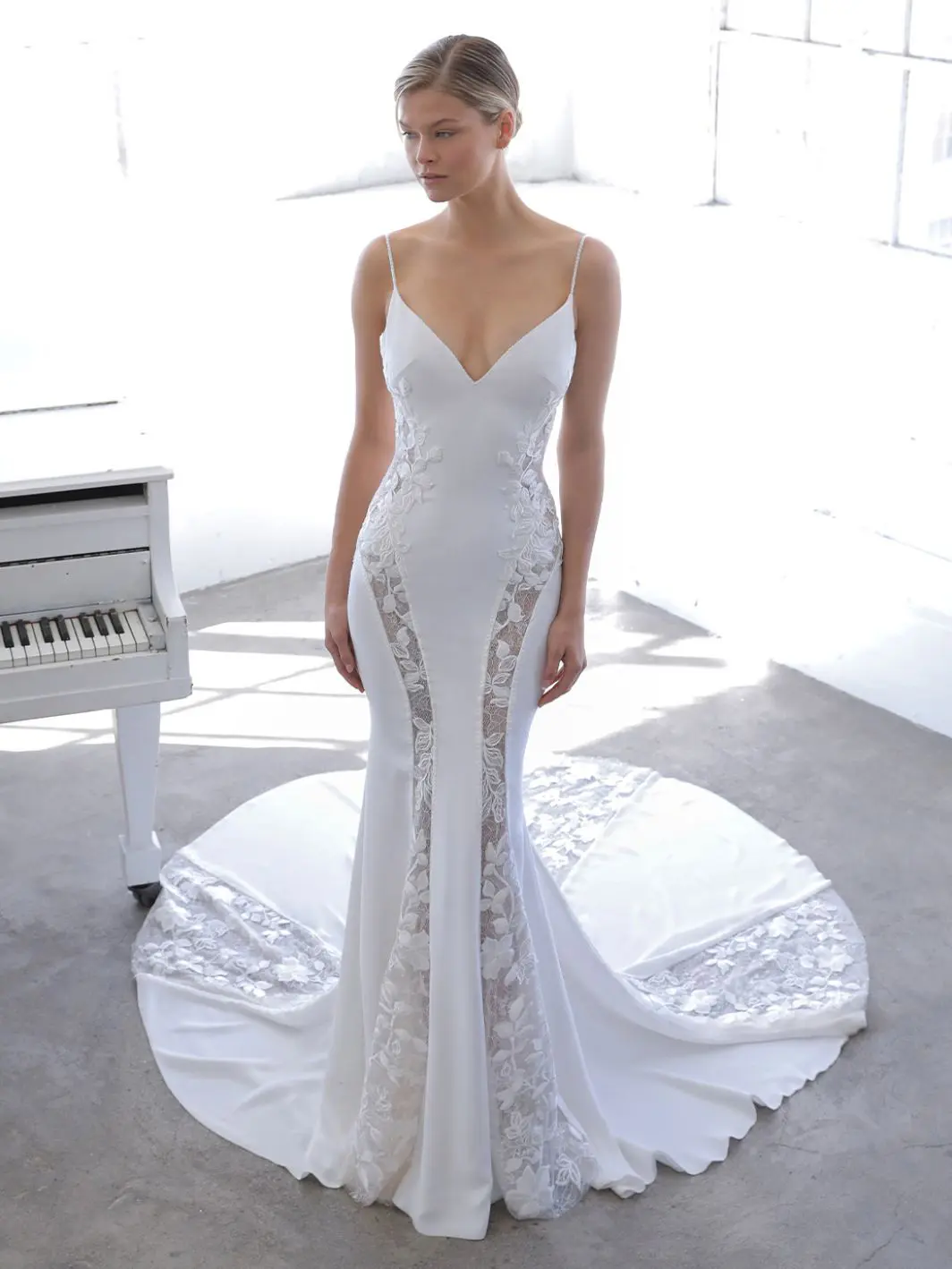 Neci, an ivory fit-and-flare bridal gown by Blue by Enzoani, features delicate spaghetti straps, a deep V-neckline, and intricate lace appliques that create sheer panels along the sides of the crepe skirt, flowing into a dramatic lace-trimmed train. The model is standing gracefully beside a white piano.