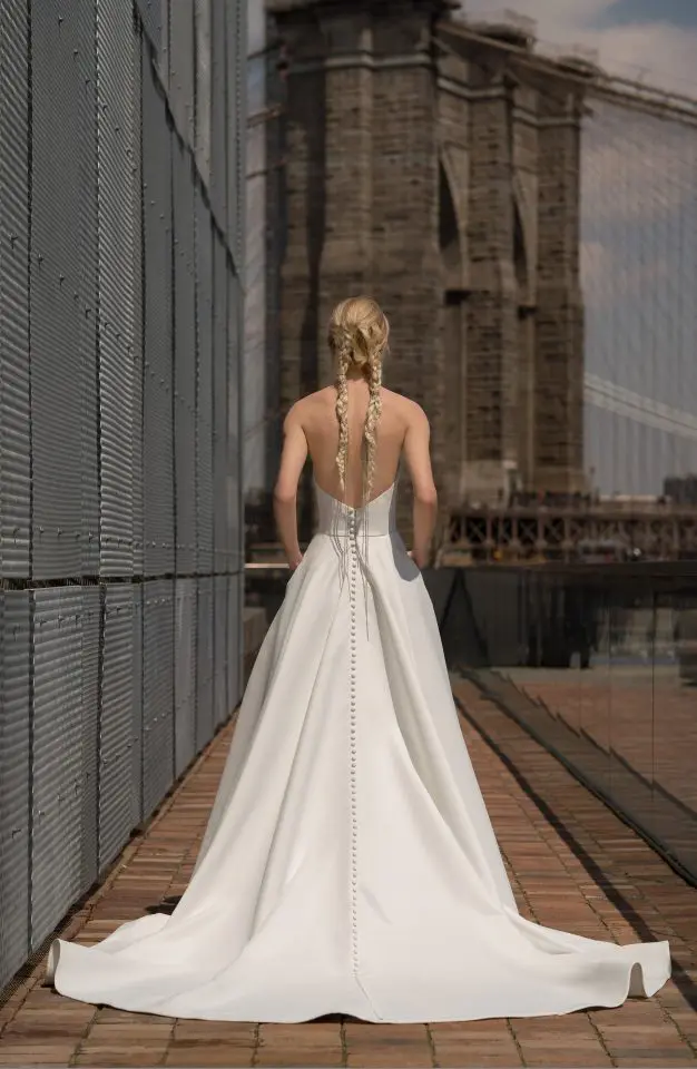 1627409467686-blanchett-back Blanchett, an elegant ivory strapless A-line bridal gown crafted from luxurious matte satin, is shown from the back. The gown features a graceful chapel-length train and a long, delicate row of buttons extending down its entire length. A blonde model, with her hair styled in two long braids, stands on a brick pathway. To her left is a corrugated metal wall, and in the sunlit background, the Brooklyn Bridge and a city skyline are visible.