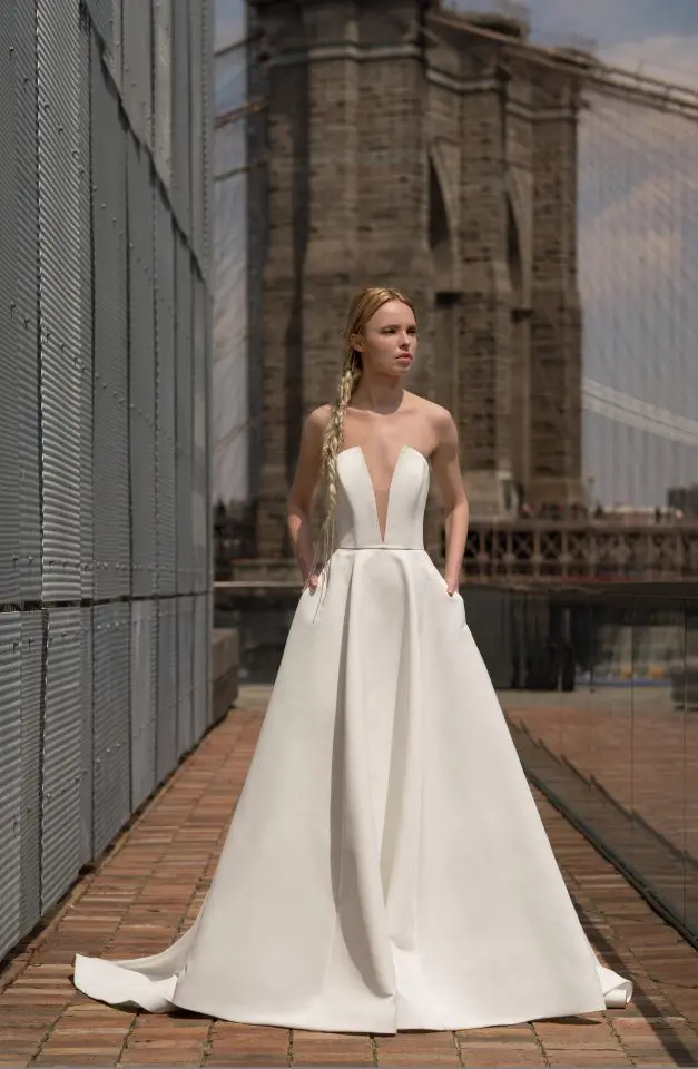 Blanchett, an ivory Alyne bridal gown with a strapless plunge V-neckline, an A-line satin skirt, and pockets, worn by a blonde model with a long braid. She stands on a brick path alongside a metal wall, with a historic stone bridge in the sunny background.