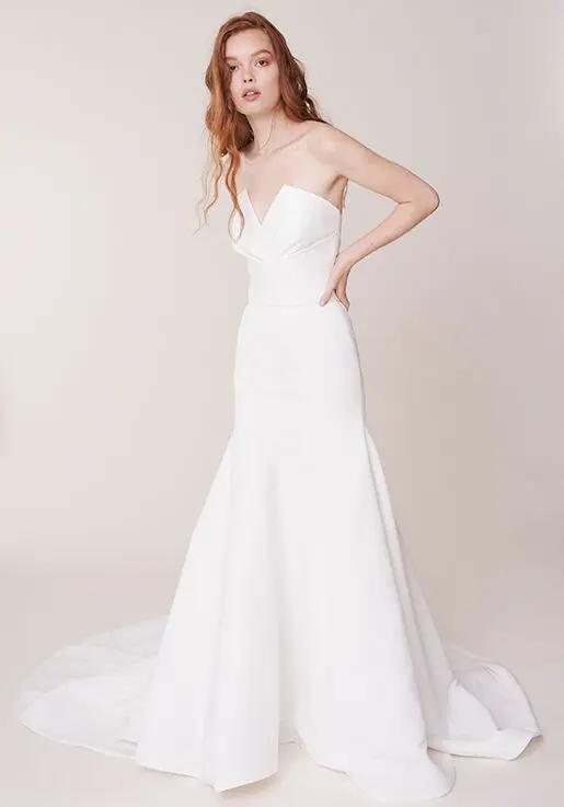 Marne, an ivory strapless mermaid bridal gown by Alyne, features a modified sweetheart neckline, a fitted satin bodice, and a chapel train. The dress is modeled by a woman with long, wavy red hair, posing with hands on her hips.