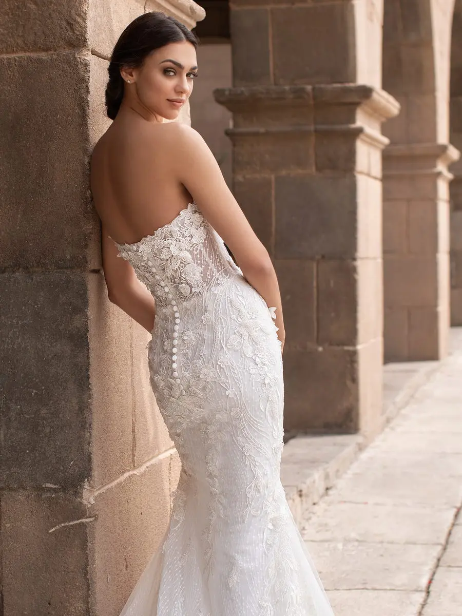 1627484778396-aethra_d_n4umj7q8xjqqtrkv Aethra, an Off White and Crystal mermaid wedding gown with a strapless sweetheart neckline, featuring intricate beaded floral appliqués over lace. The fitted bodice reveals a delicate row of buttons down the back, hugging the body and hips before flaring into a dramatic skirt. A bride with dark hair styled in a low bun leans casually against an old stone wall, looking back over her shoulder with a subtle smile, in an outdoor setting with stone arches in the background.