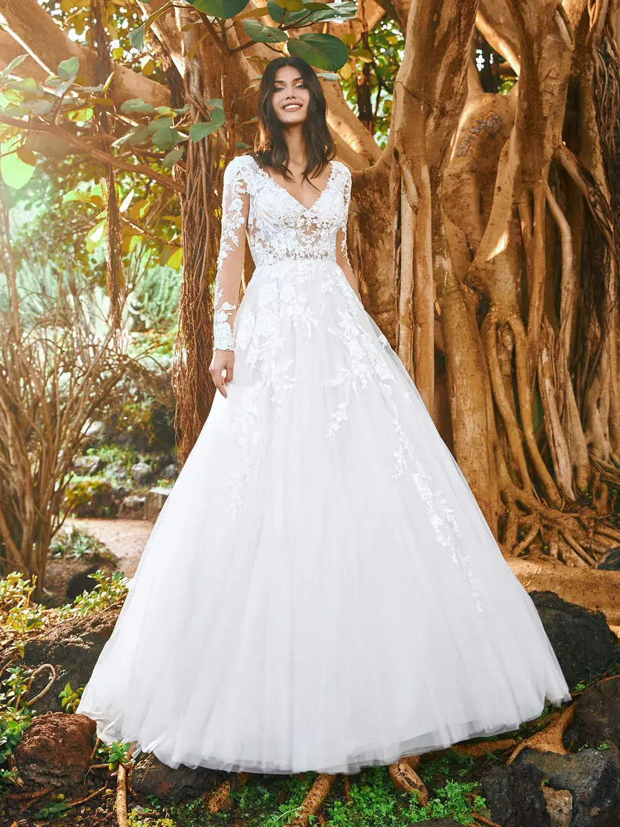 Maroon, an off-white Pronovias A-line princess bridal gown, features a V-neck illusion bodice and long sheer sleeves, both intricately adorned with lace and embroidery. The gown flows into a voluminous tulle skirt, worn by a smiling model posing outdoors amidst large tree trunks and roots.