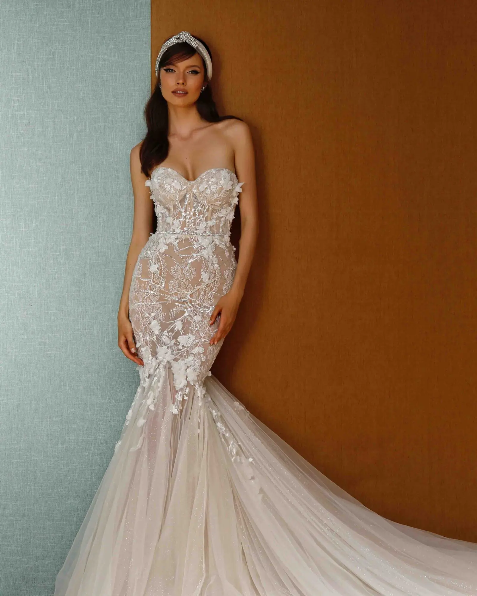 21-P02, a Berta Privee ivory and nude mermaid bridal gown, featuring a strapless sweetheart corset bodice intricately adorned with lace, sparkling beading, and 3D floral appliques. The fitted silhouette extends into a shimmering chapel-length tulle train. The model wears a jeweled headband and poses against a light blue and brown textured wall.