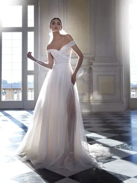 Ida, an off-white and light beige Pronovias bridal gown, features an off-the-shoulder V-neck bodice and sheer long sleeves, both shimmering with embellishments. The Fit n Flare silhouette flows into a voluminous tulle skirt with a high slit and a cathedral train. The model elegantly poses in a sunlit, opulent hall.