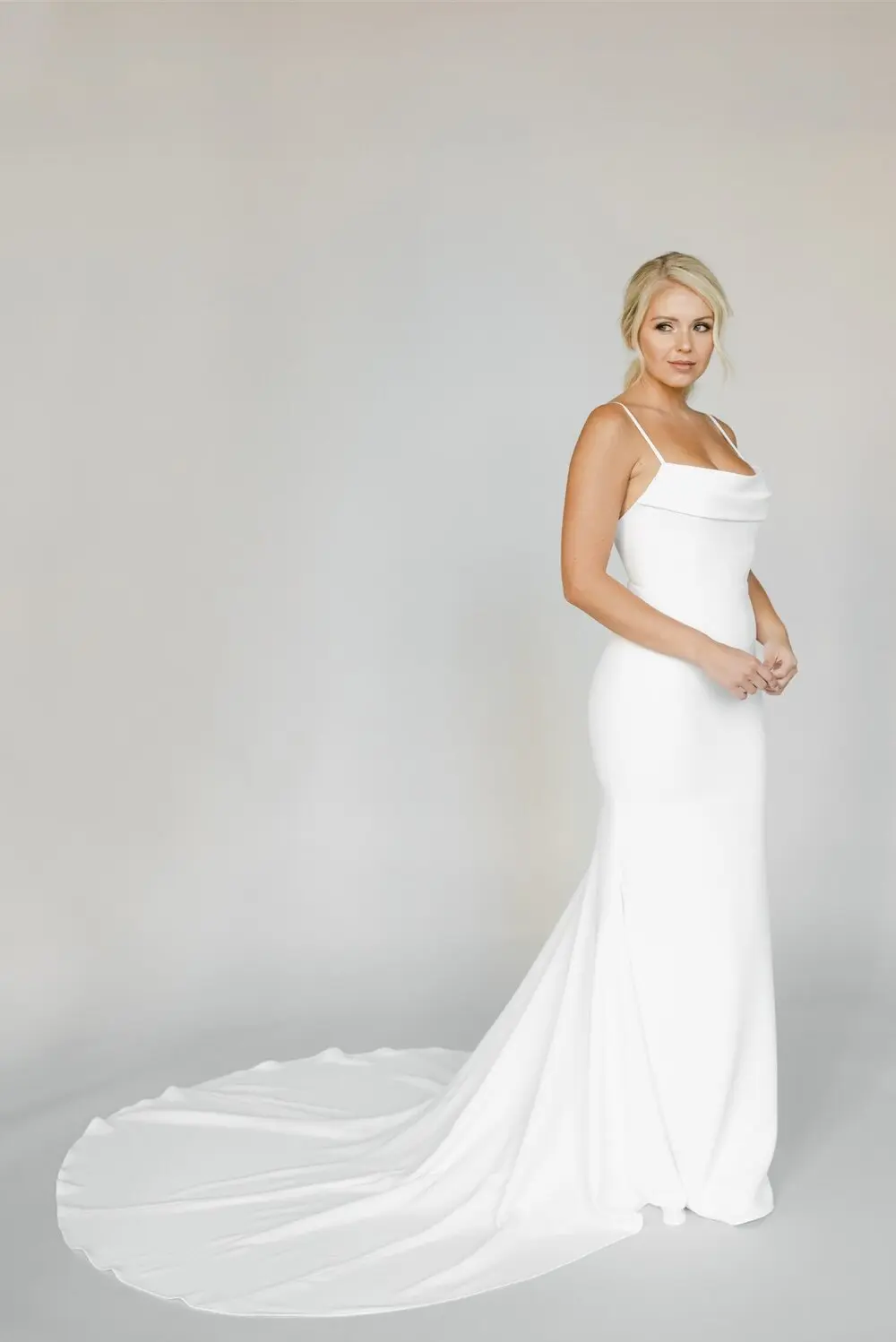 Esme, an ivory fit and flare bridal gown by Alyssa Kristin, featuring spaghetti straps, a scoop neckline, a chapel length train, and made of crepe fabric. The gown is modeled by a blonde woman looking to her right, standing against a light grey background.