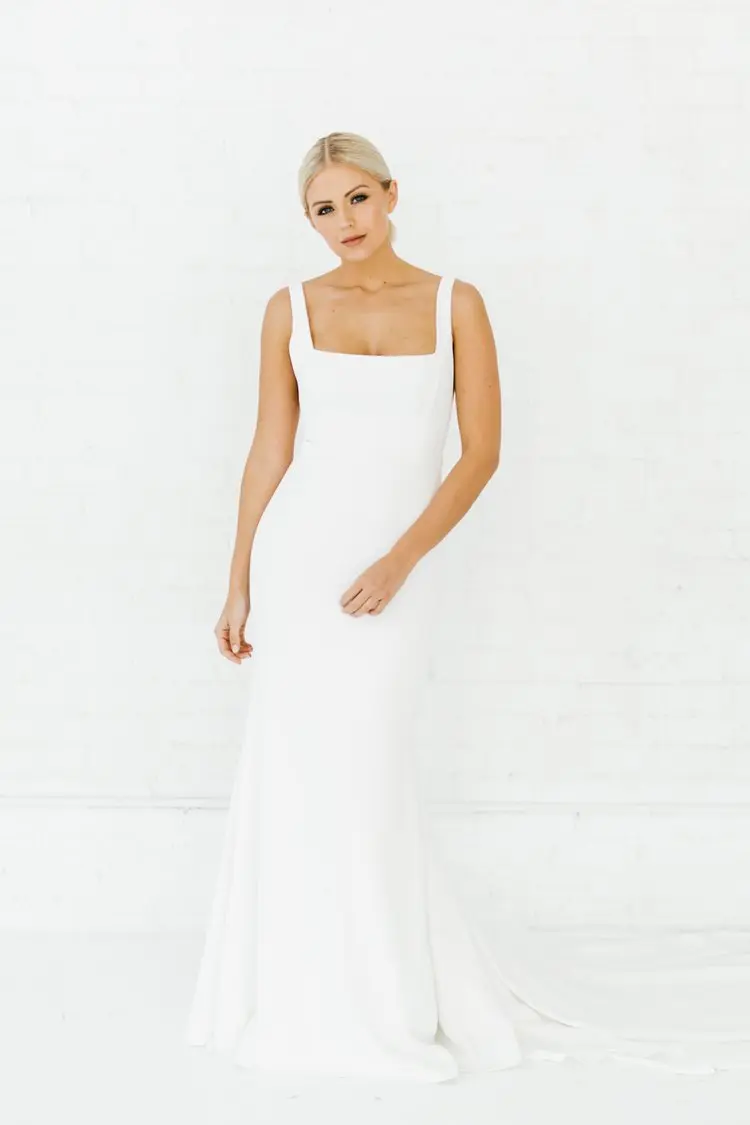 Sydney, an ivory Alyssa Kristin bridal gown with a square neckline, tank straps, and a sleek fit-and-flare silhouette in crepe fabric. The floor-length dress features a subtle train and is worn by a blonde model posing against a white brick wall.