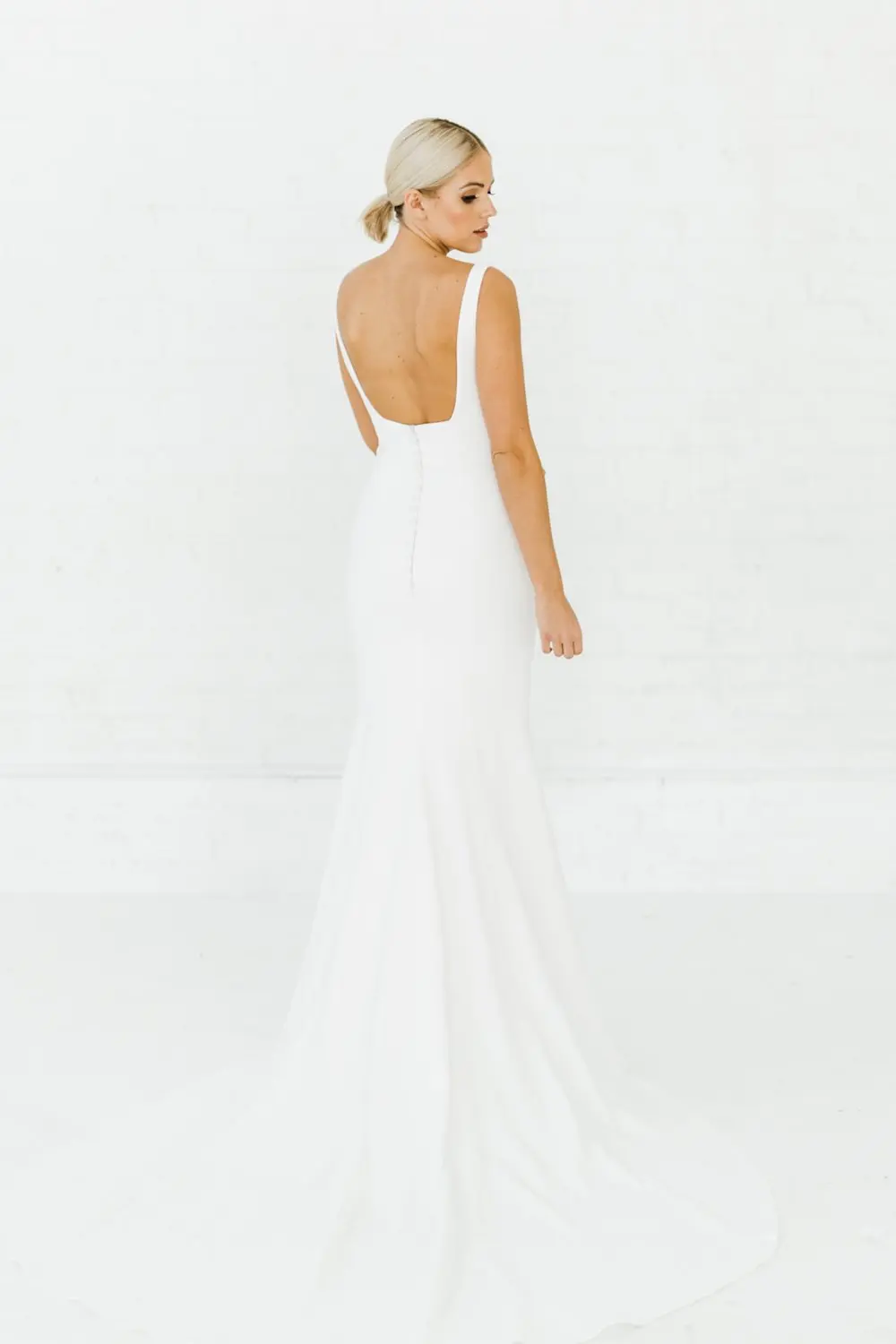 1698352635099-ARP20229 Sydney, an ivory Alyssa Kristin fit-and-flare bridal gown, is worn by a blonde model facing away, looking over her right shoulder. The gown features a deep U-back, thin tank straps, and a line of buttons down the back. Made from sculpting stretch crepe, it hugs the figure with subtle nude mesh side cutouts and flares into a graceful sweep train.