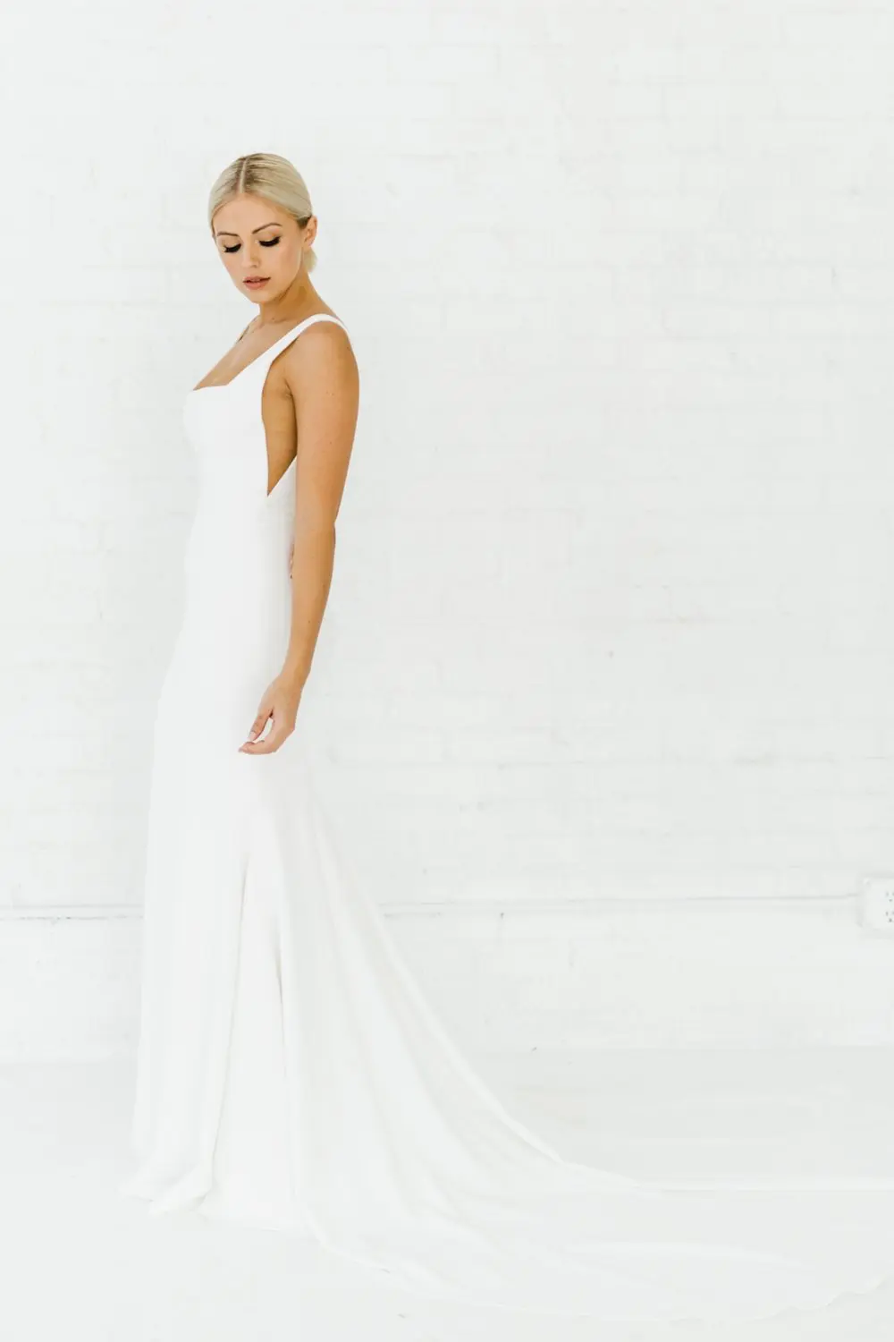 1698352635102-ARP20184 Sydney, an ivory Alyssa Kristin bridal gown, features a sleek U-neckline and subtle nude mesh side cutouts. Crafted from signature stretch crepe, this fit-and-flare silhouette smooths and sculpts the figure, flowing into a graceful floor-length skirt with a train. A blonde model with an elegant updo poses in profile against a white brick wall.
