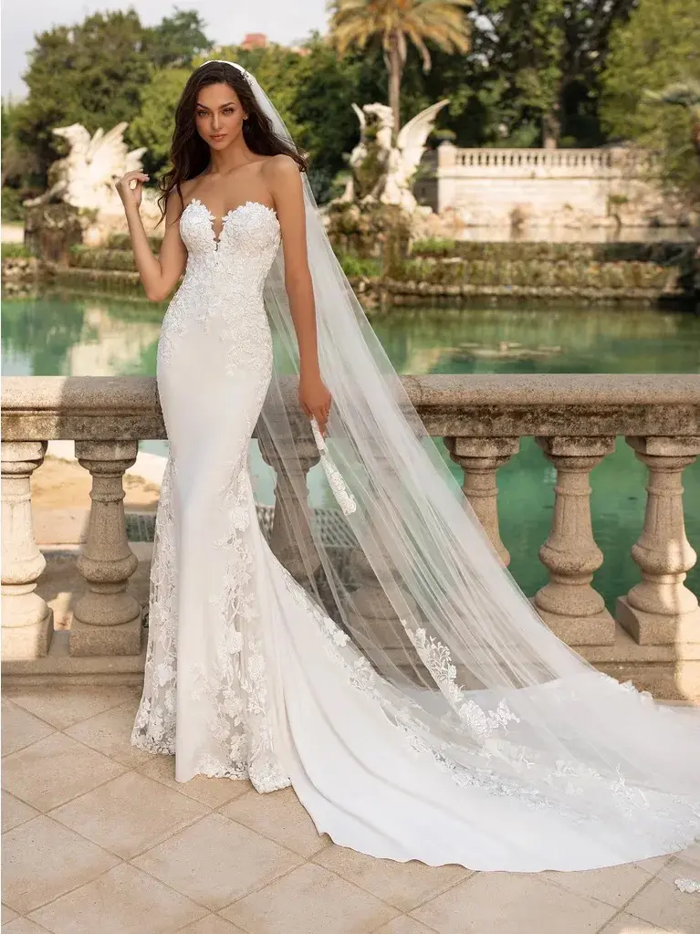 Epico, an off-white and crystal mermaid wedding gown by Pronovias, features a strapless sweetheart neckline and is adorned with delicate lace applique, embroidery, and beading over crepe and tulle, with a chapel train. The bride wears a long, lace-trimmed veil and holds a white fan while posing on a stone balustrade overlooking a picturesque garden pond with classical sculptures.