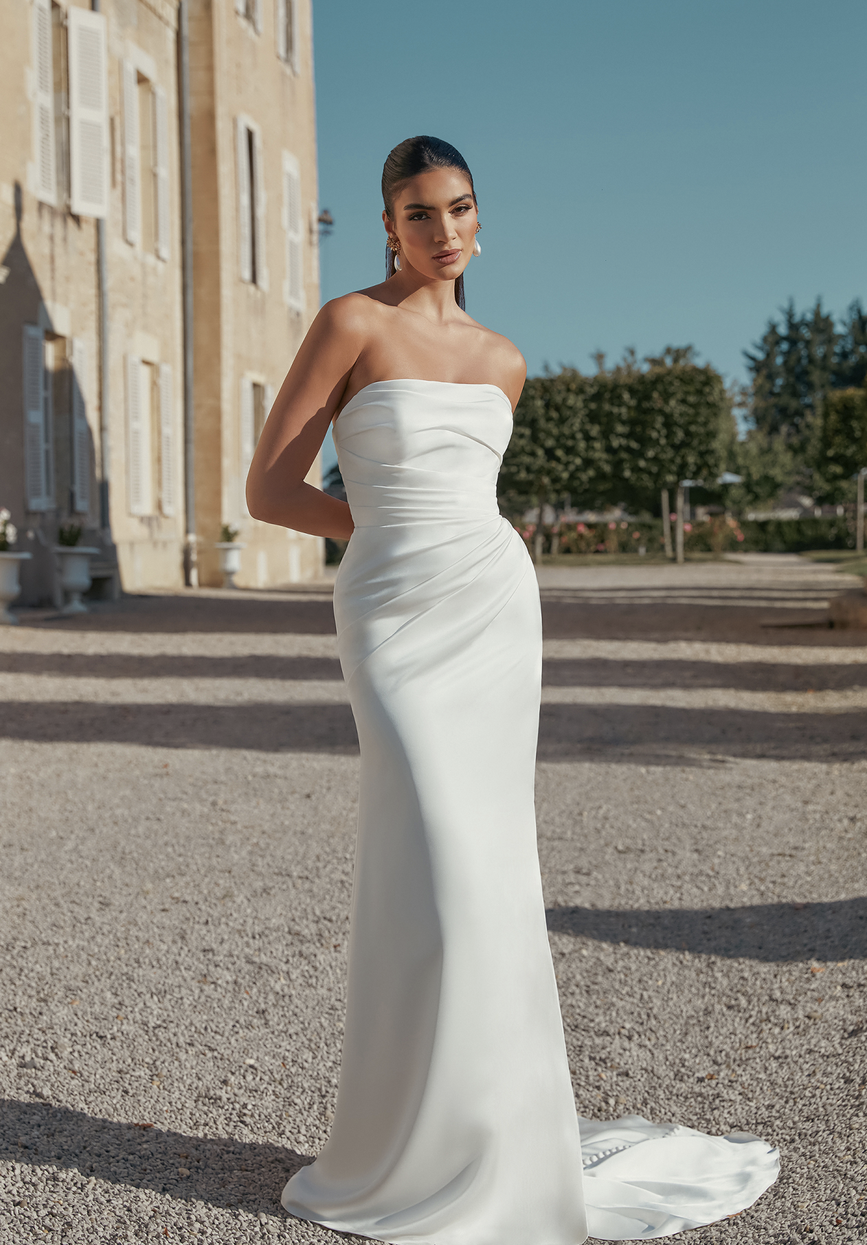 An ivory strapless Charmeuse fit-and-flare bridal gown by Sincerity by Justin Alexander, with ruching on the bodice and hip, and a sweep train featuring buttons. A model wearing the gown and ornate earrings stands outdoors on a gravel path, with a historic building and garden under a blue sky.