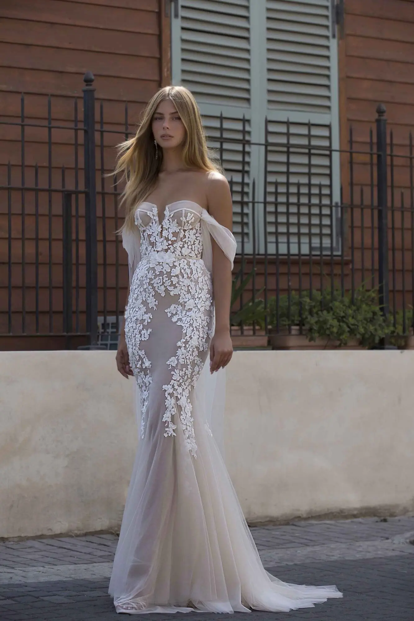 1729888090984-image-33 21-P106, a stunning nude and ivory lace mermaid wedding gown adorned with delicate floral appliqués. It features a corset-style strapless sweetheart bodice with a slightly plunging neckline and elegant off-shoulder tulle sleeves that cascade softly toward a dramatic train.