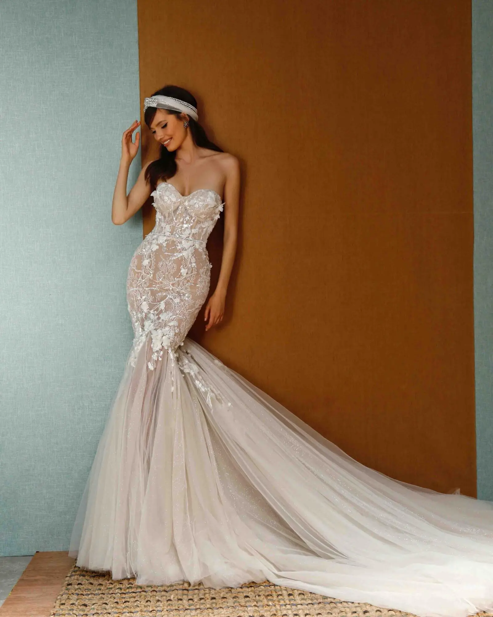 1729889451183-image-39 A radiant Berta Privee strapless mermaid wedding gown, featuring a nude base with intricate ivory lace appliqués and sparkling beading. The dress has a sweetheart neckline, a corseted bodice that sculpts the silhouette, and a dramatic, flowing tulle train. The model, smiling softly and wearing a delicate beaded headband, poses against a textured blue and brown wall.