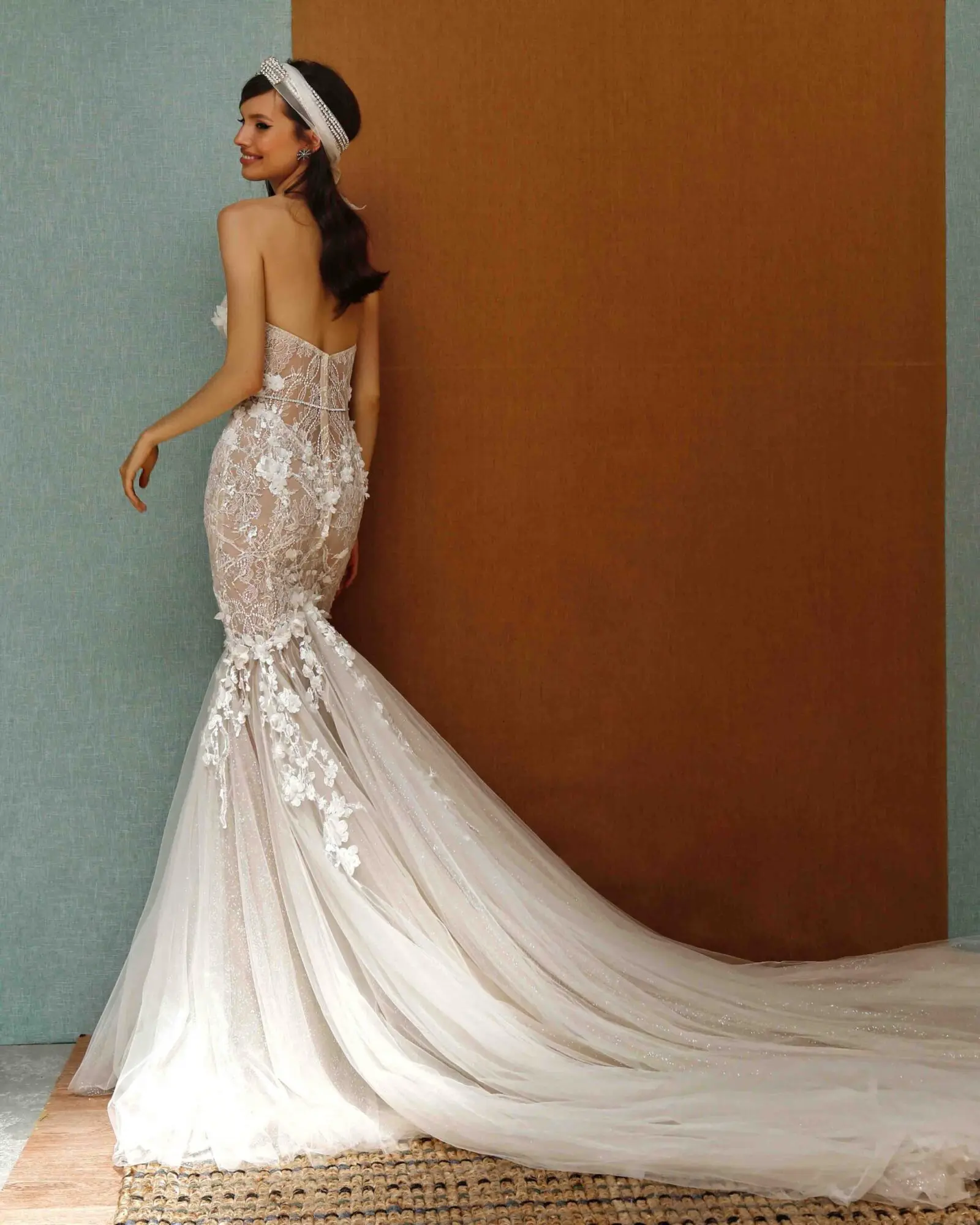 1729889451184-image-40 21-P02, an ivory and nude strapless corseted mermaid bridal gown by Berta Privee. Adorned with intricate lace appliqués and subtle beading, the dress features a sweetheart neckline, a sculpted bodice, and a dramatic chapel-length tulle mermaid train. A smiling model, wearing a jeweled headband, looks back over her right shoulder, posing against a textured wall of blue-green and brown.