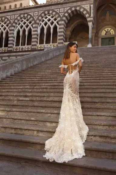 1730231042597-image-11 20-P105, an AS SAMPLED (nude and ivory) Berta Privee fit and flare bridal gown, features a corset strapless sweetheart bodice with off-shoulder straps and an open lace-up back. Intricate floral lace appliqués and delicate beading adorn the sheer fabric, cascading from the bodice down the flowing skirt to a short train. A model, with long brown hair, stands on wide stone steps, smiling over her shoulder, with a grand building featuring striped arches in the background.
