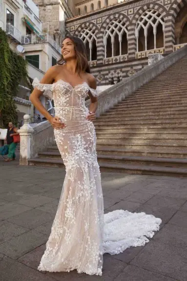 1730231042598-image-10 A stunning Berta Privee bridal gown with a nude-colored corset, strapless, fit and flare silhouette, and delicate off-shoulder straps framing a sweetheart neckline. The dress is intricately designed with white floral lace appliqués and subtle beading that cascade down the sheer fabric, ending in a graceful train. A smiling model with long brown hair poses outdoors on a stone path, with a grand building featuring striped arches and wide stone steps in the background, suggesting an elegant Mediterranean or European setting.