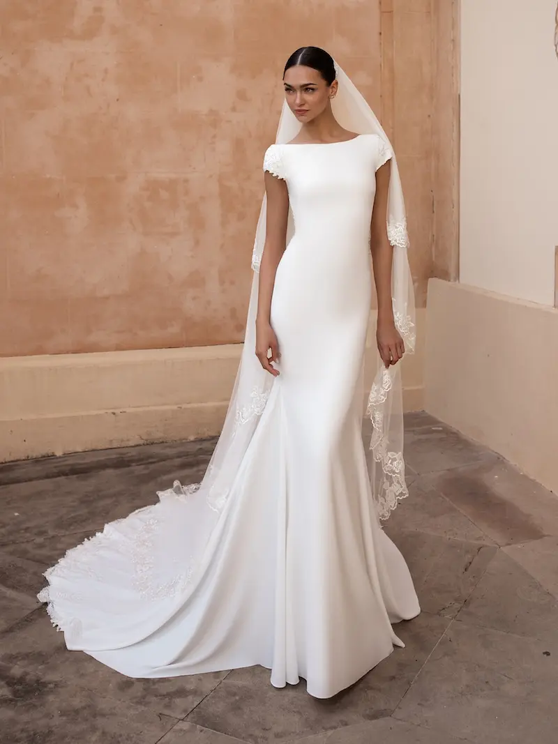 ANITRA, an off-white Pronovias bridal gown featuring a fit-and-flare silhouette, a modest bateau neckline, and delicate cap sleeves. The crepe gown showcases a chapel-length train adorned with intricate lace and embroidery, complemented by a matching lace-edged veil.